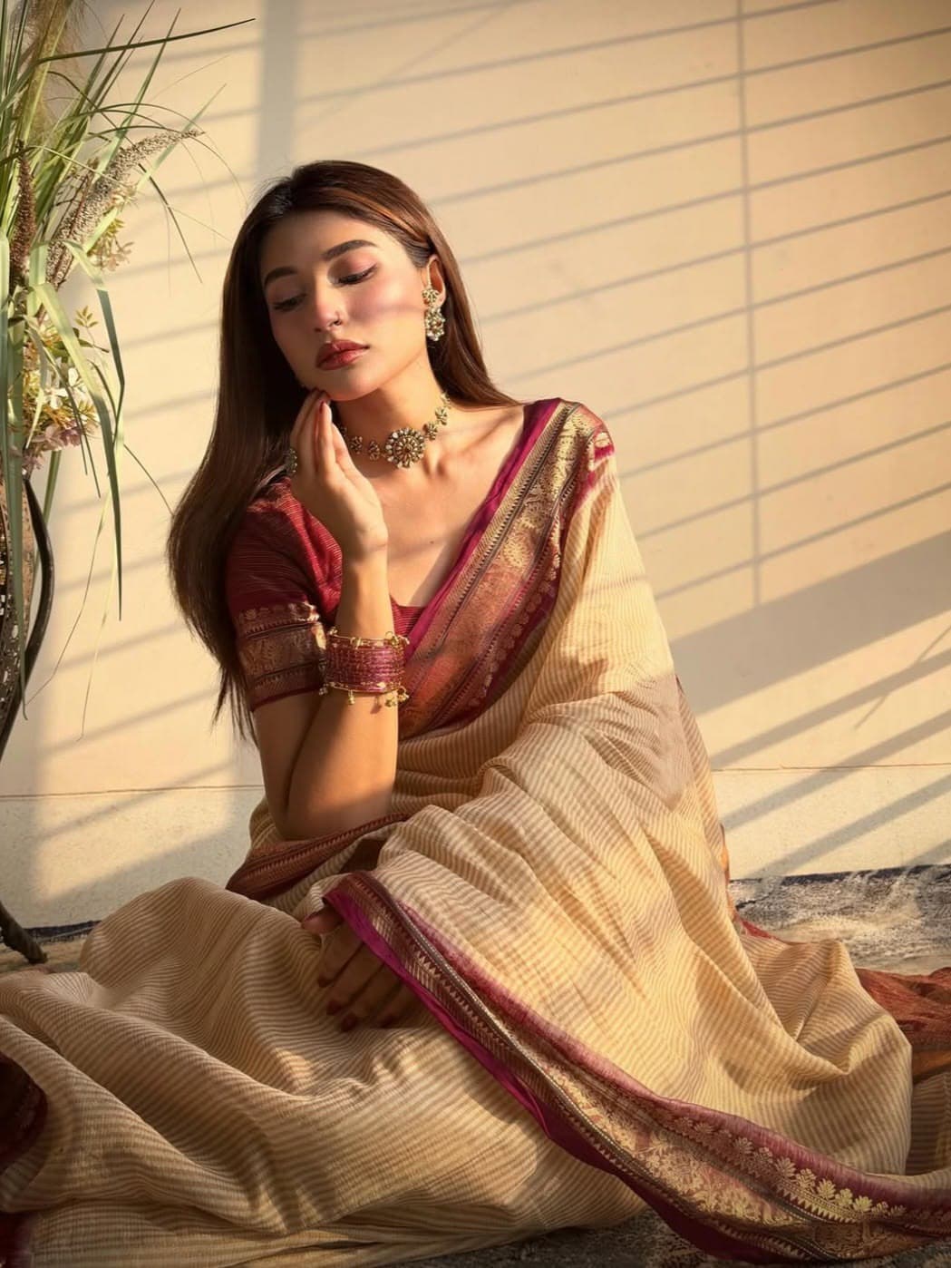 Woman seated against a sunlit wall with louvred shadow lines, wearing a cream and maroon banarasi saree